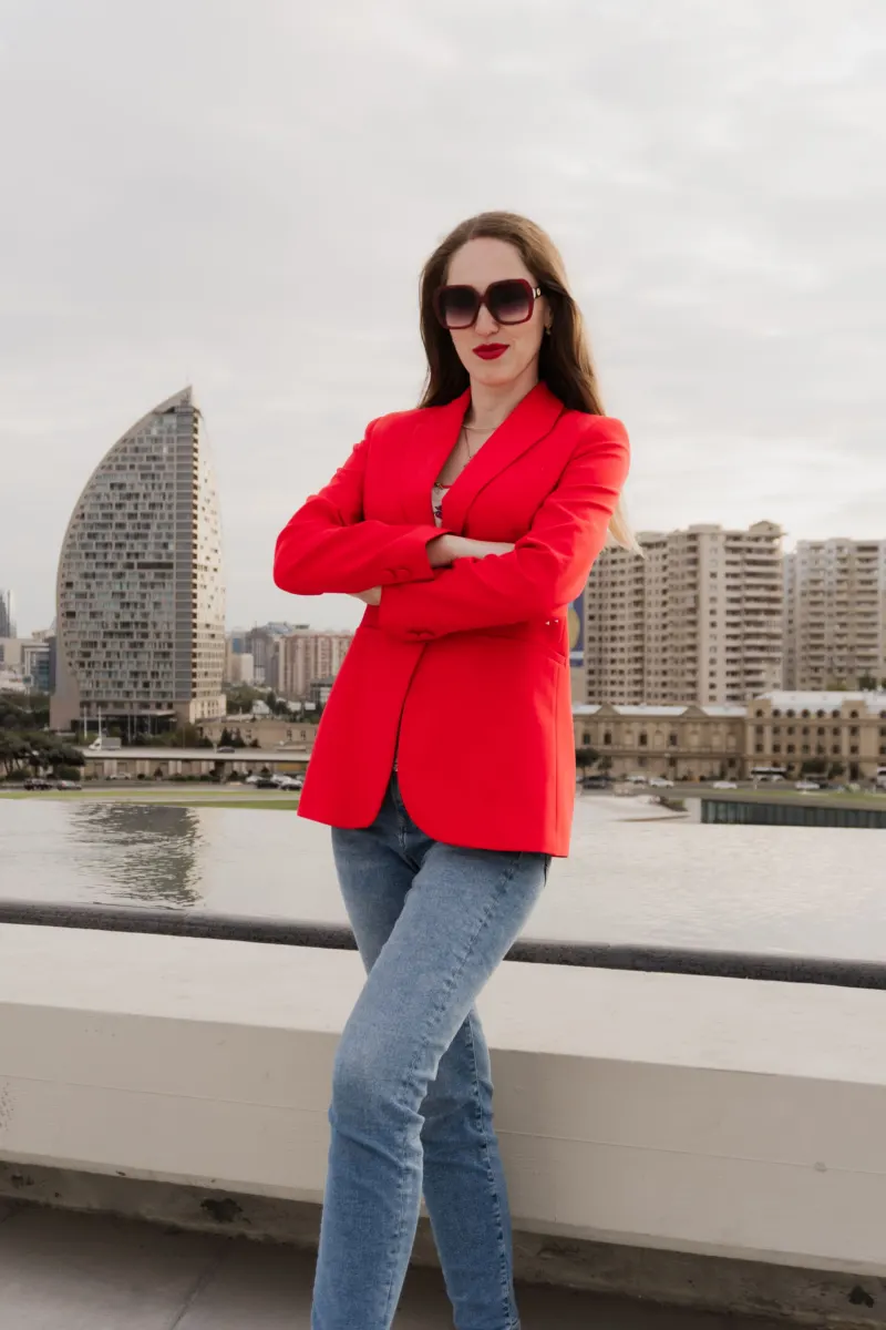 Woman in a red blazer, blue jeans and dark sunglasses on a waterfront terrace