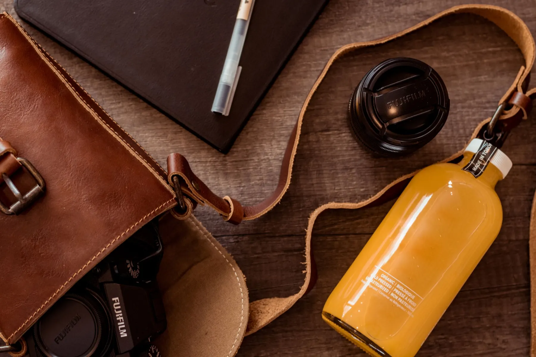 Open brown leather bag on a table with a notebook, lens cap and bottle beside it