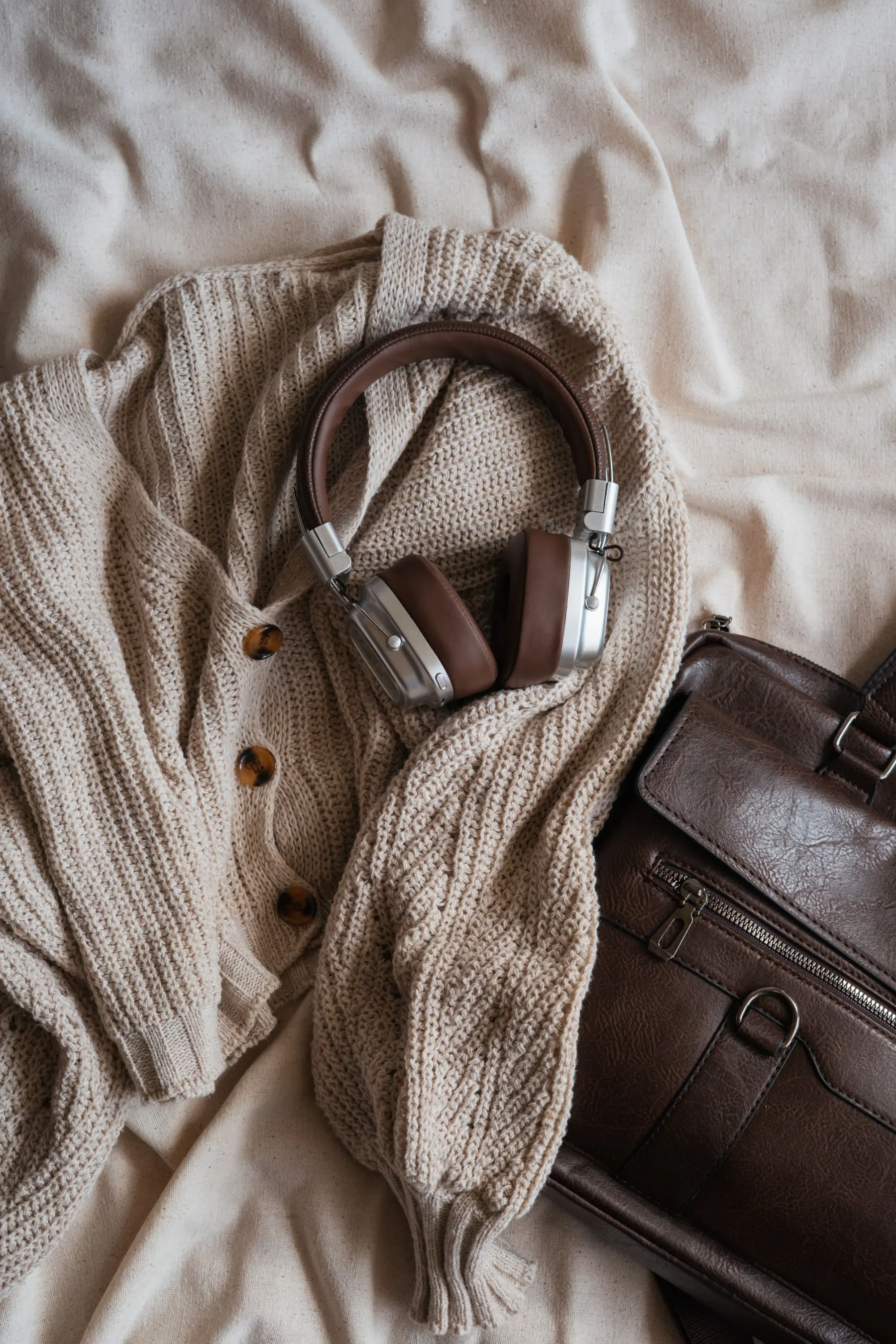 Cream cable-knit cardigan, brown leather bag and headphones on a linen sheet