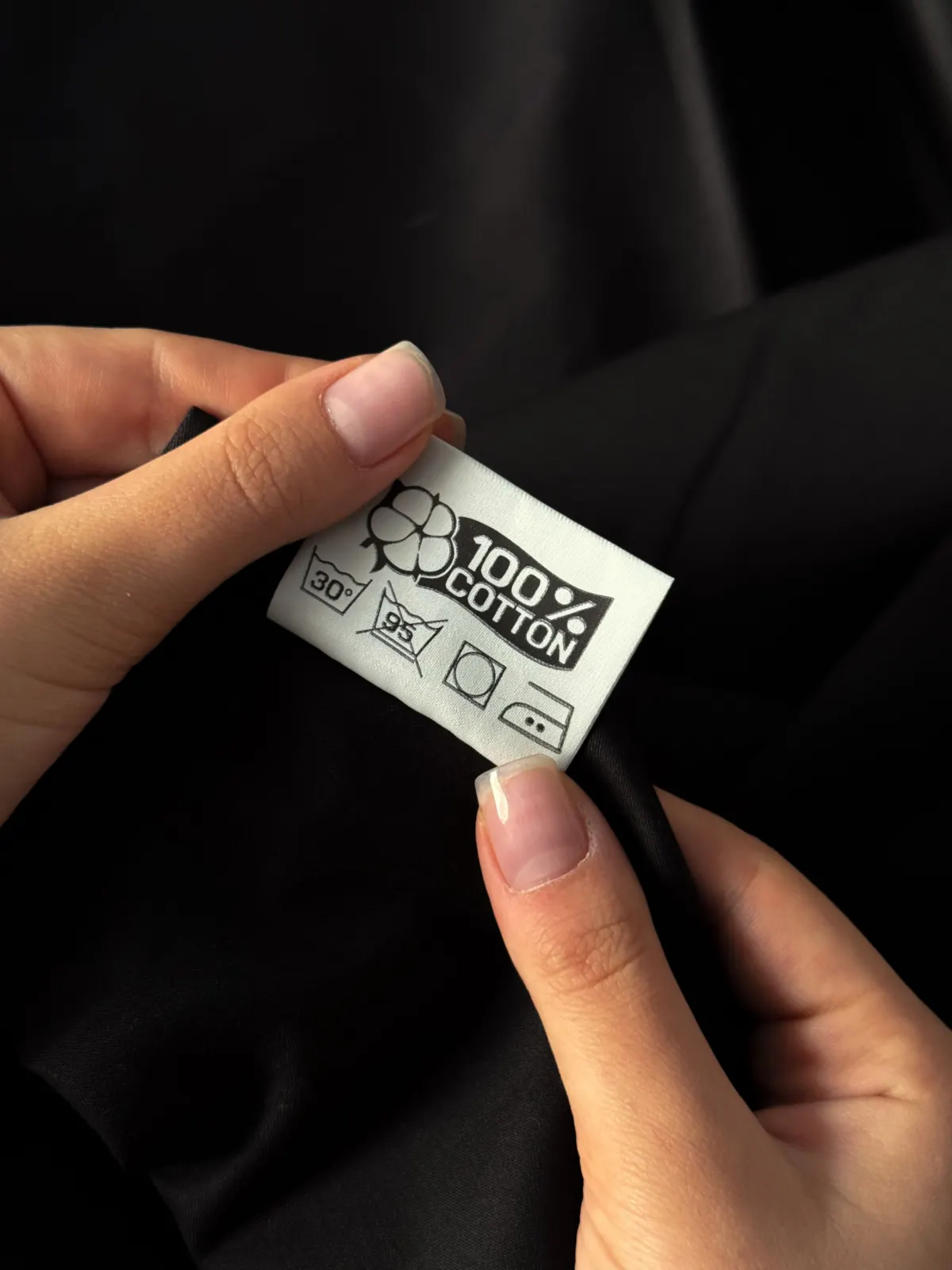 Fingers holding a sewn-in care label that reads 100% cotton above a row of care symbols