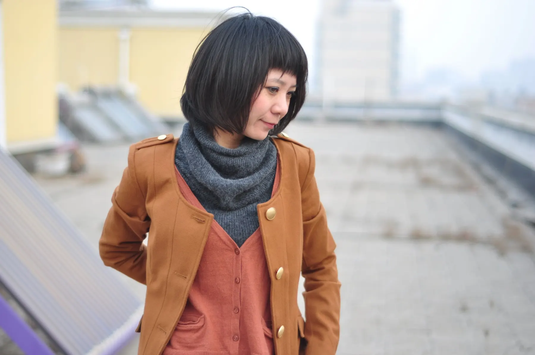 Woman in a rust jacket over a shirt with a grey snood, standing on a rooftop