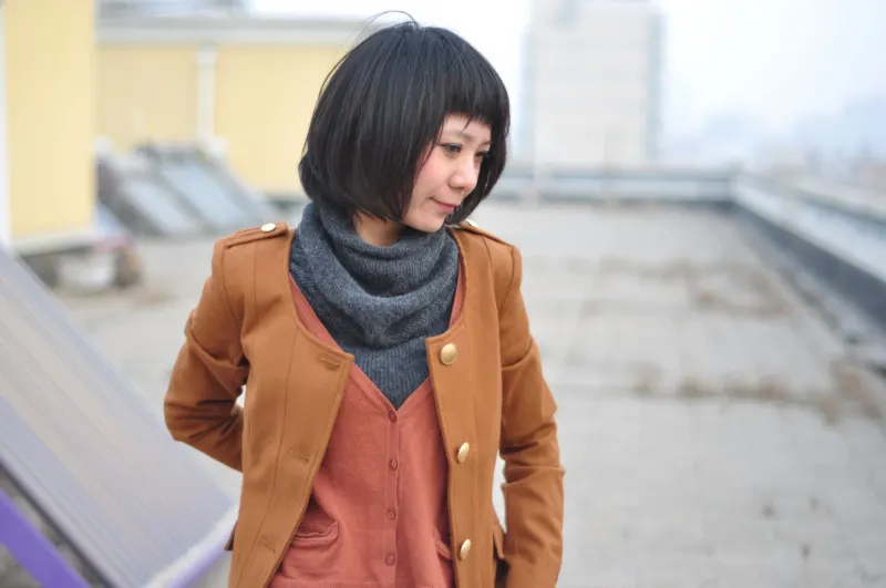Woman in a rust jacket over a shirt with a grey snood, standing on a rooftop