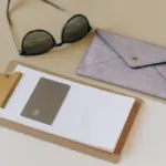 Sunglasses, a clipboard and a grey wallet on a beige surface