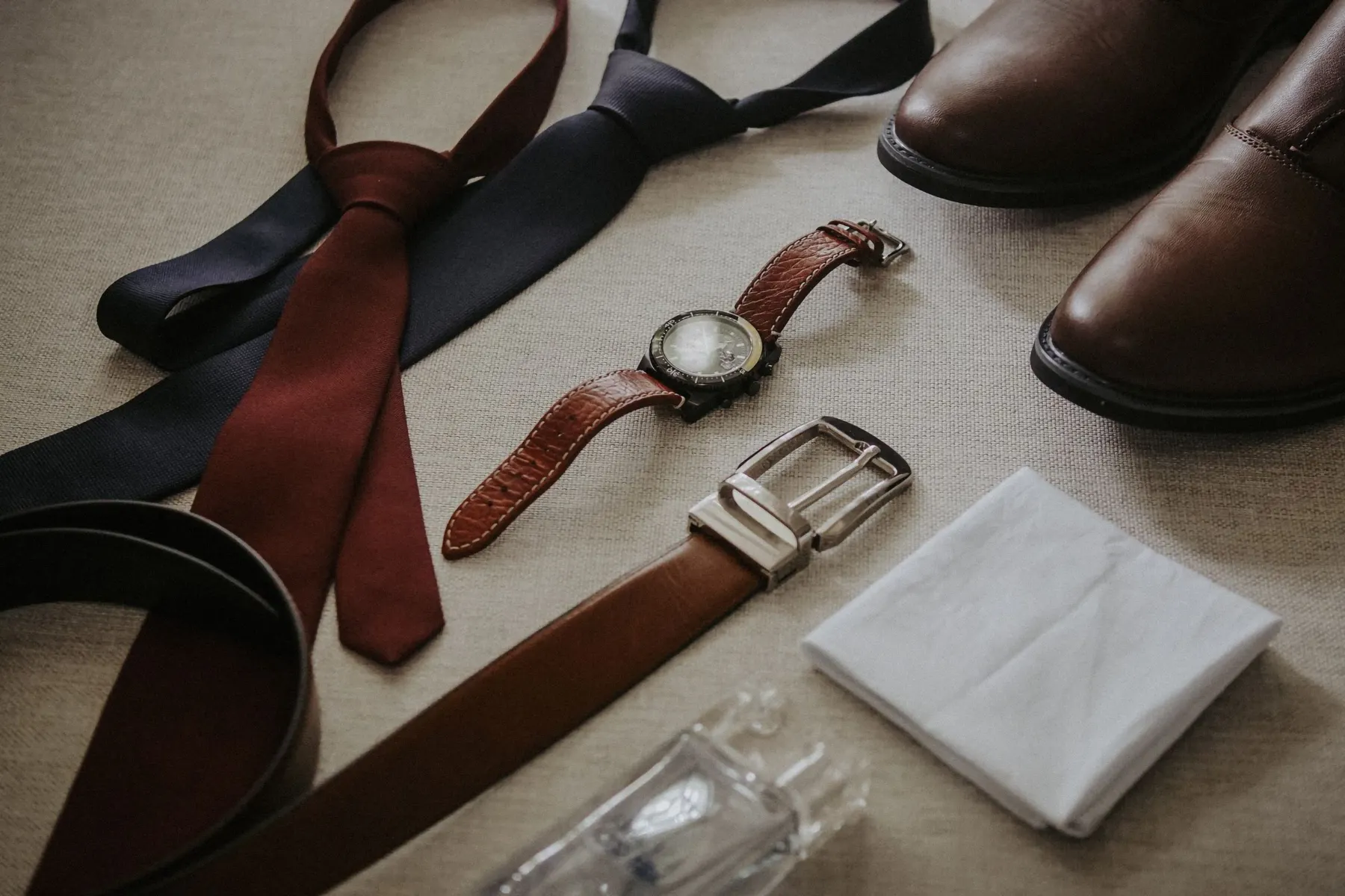 Leather belts, ties, a watch, a pocket square and brown shoes laid out on a table