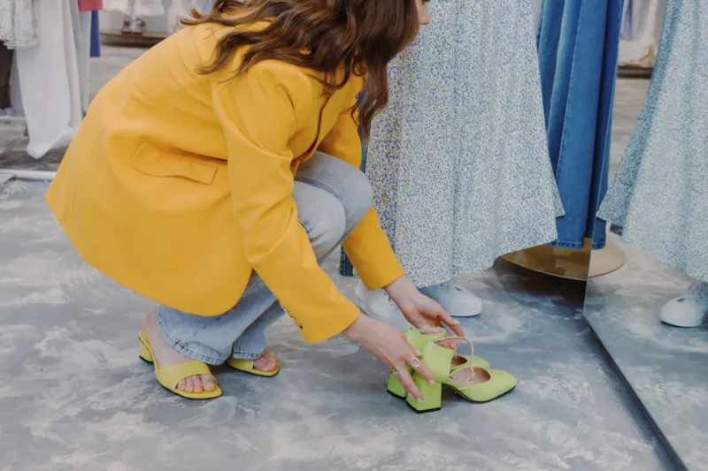 Woman in a yellow jacket crouching to try on a pair of green heeled sandals