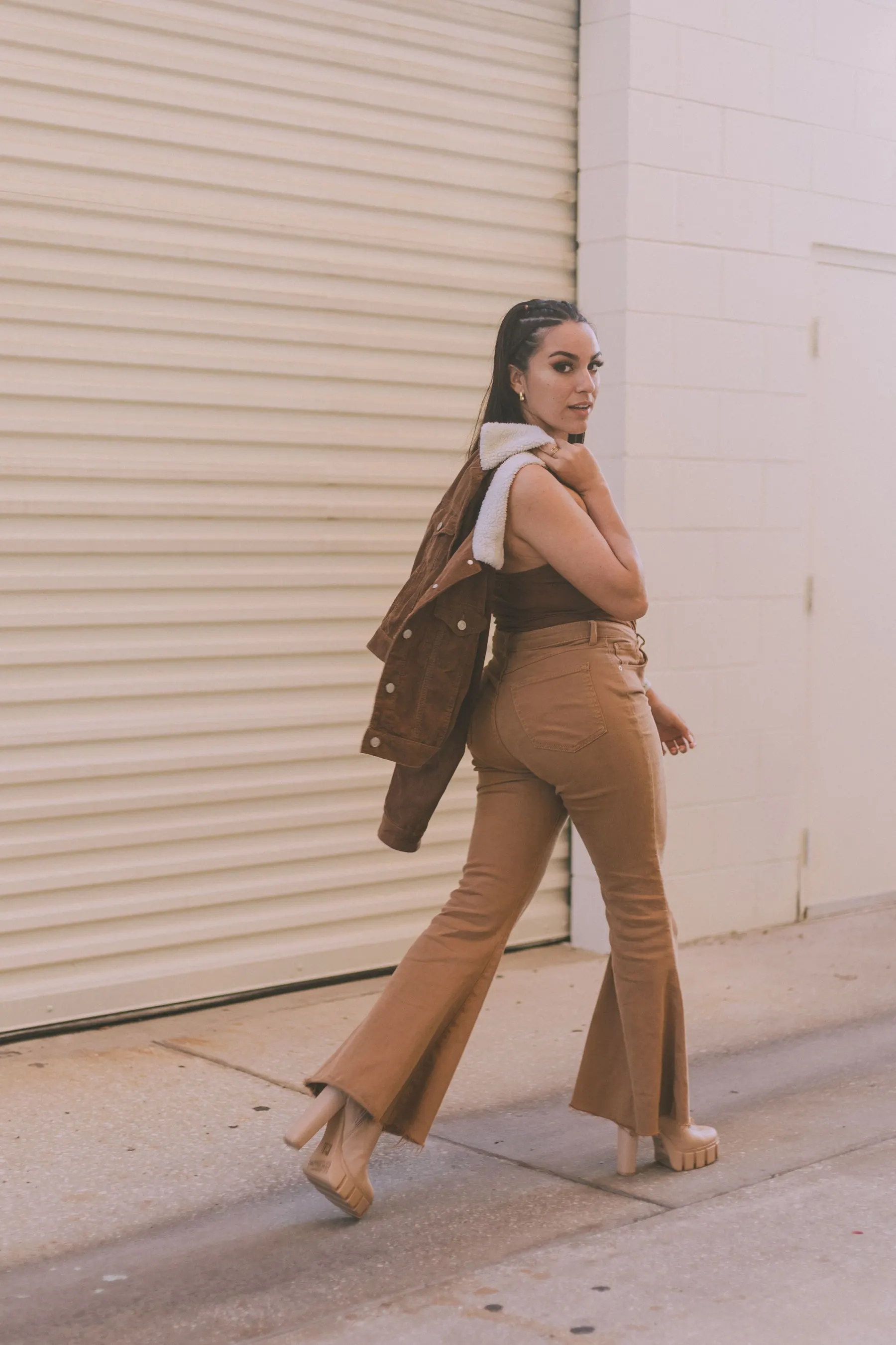 Woman in tan flared trousers and a brown top with a jacket over her shoulder