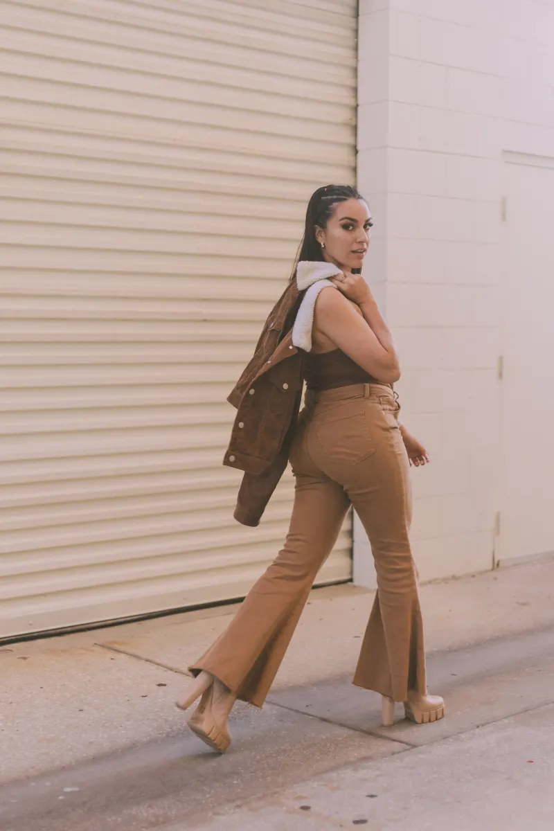 Woman in tan flared trousers and a brown top with a jacket over her shoulder