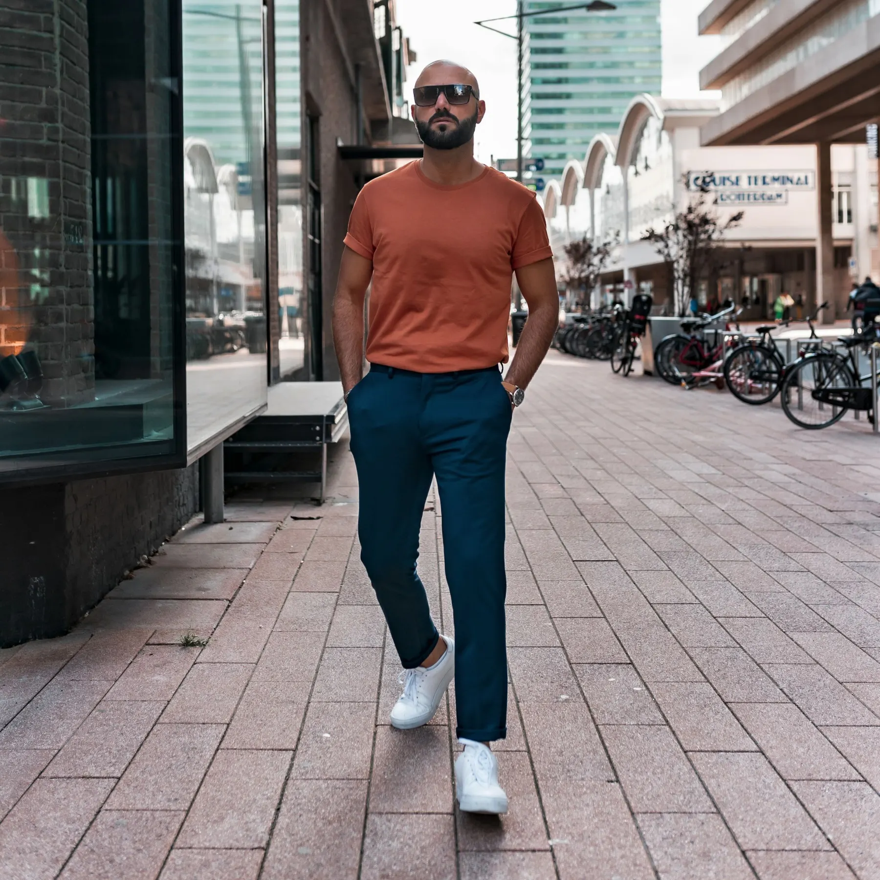 Man in a rust T-shirt, navy trousers and white trainers walking along a street