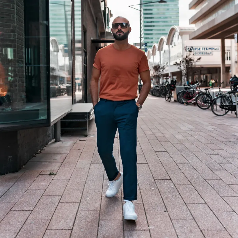 Man in a rust T-shirt, navy trousers and white trainers walking along a street