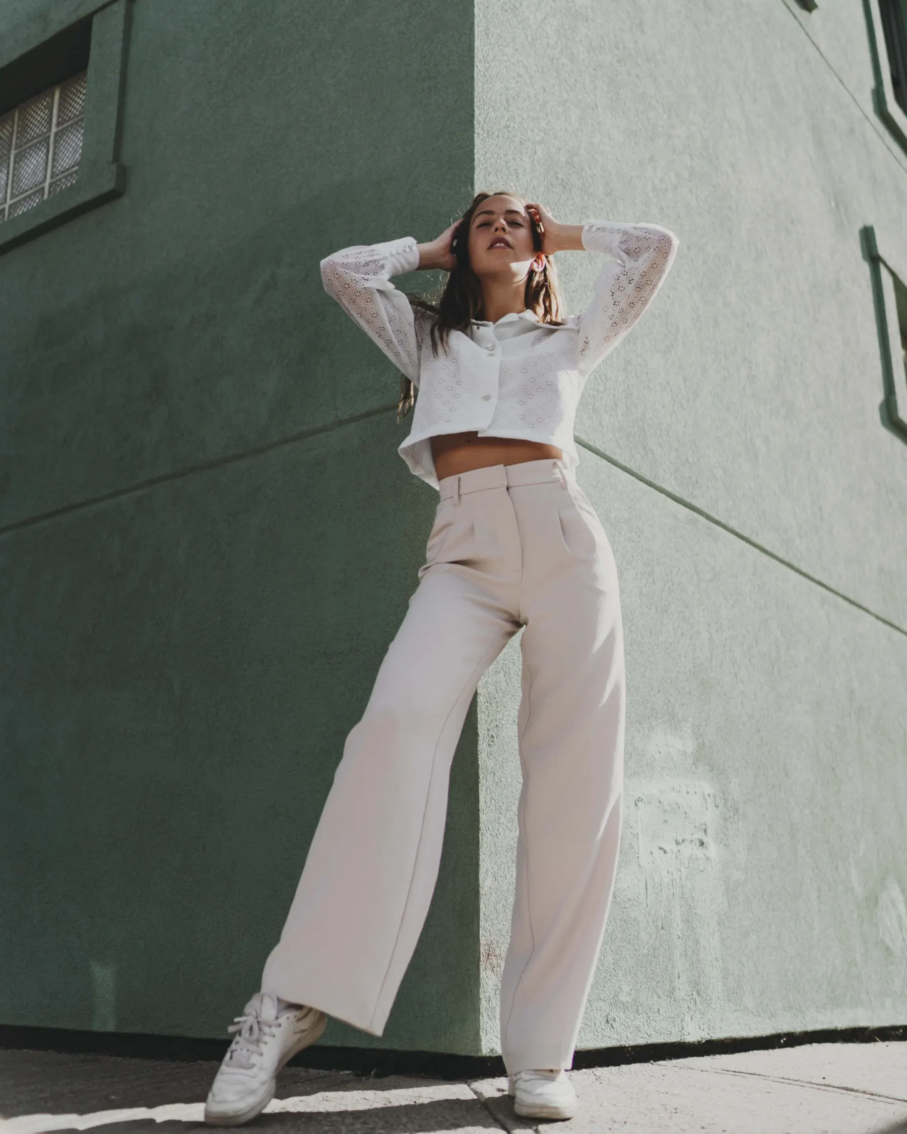 Woman in wide cream trousers, a white cropped shirt and white trainers by a green wall